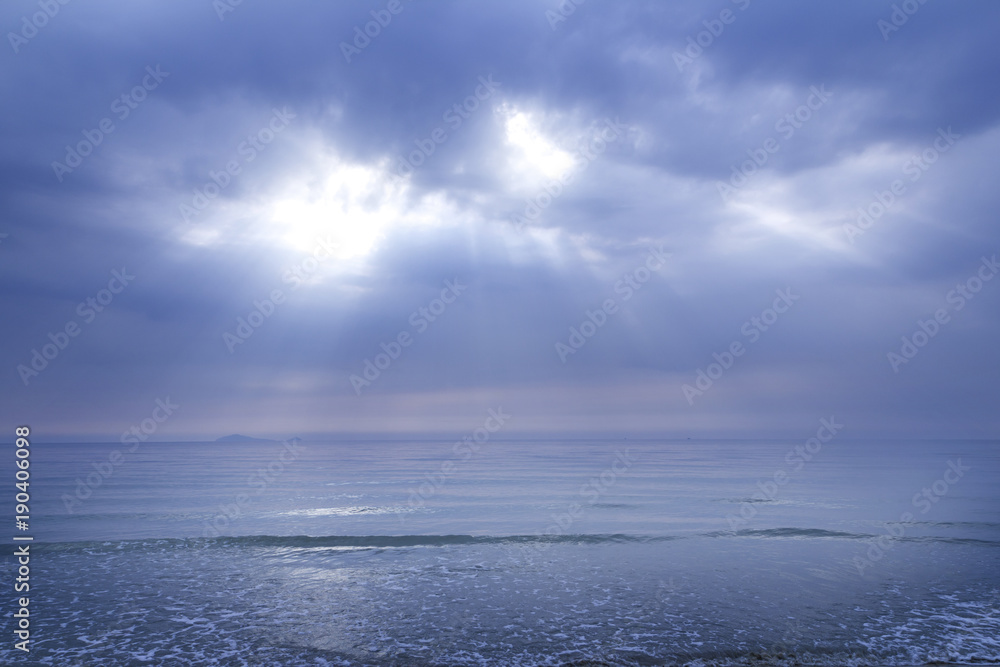 Obraz premium Sun shining through thick clouds of overcast cloudy sky caused ray of light beaming down to the blue ocean surface