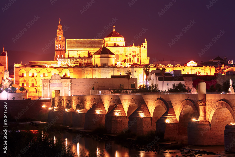 Illuminated Great Mosque Mezquita - Catedral de Cordoba with mirror reflection and Roman bridge across Guadalquivir river during evening blue hour, Cordoba, Andalusia, Spain
