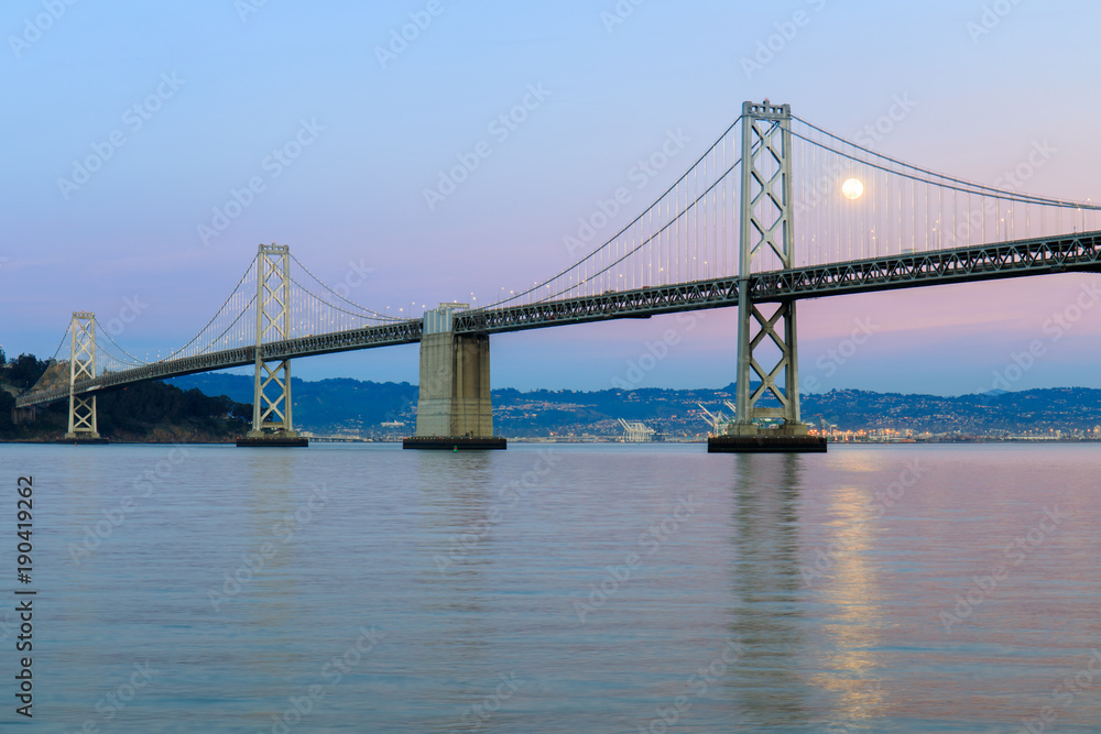 San Francisco-Oakland Bay Bridge with full moon rising and pink and blue sunset skies. The Embarcadero, San Francisco, California, USA.