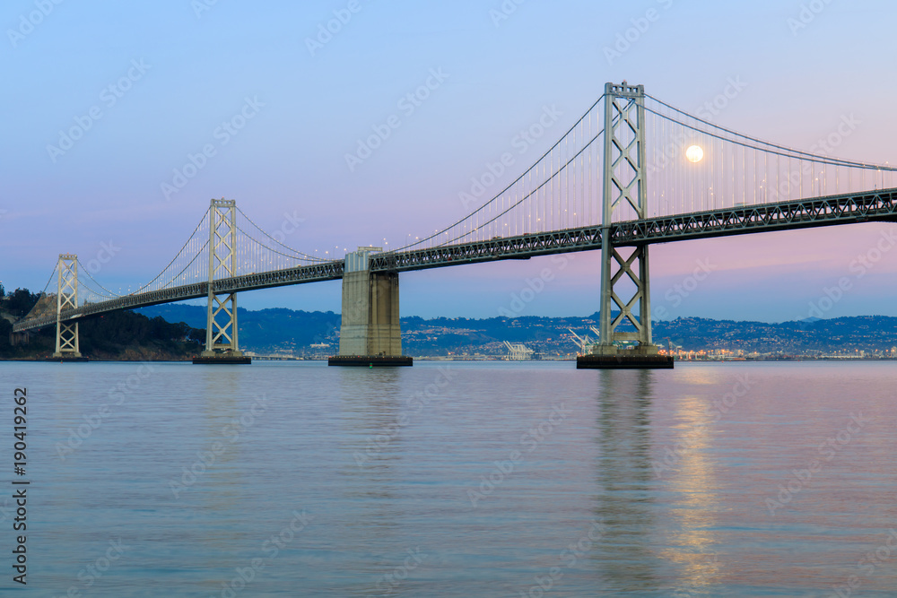 San Francisco-Oakland Bay Bridge with full moon rising and pink and blue sunset skies. The Embarcadero, San Francisco, California, USA. San Francisco-Oakland Bay Bridge with full moon rising and pink and blue sunset skies. The Embarcadero, San Francisco, California, USA.