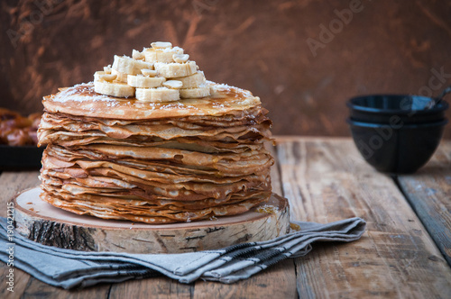 On the table on a stand made of wood cut a pile of thin pancakes, sprinkled with jam, sprinkled with powdered sugar, on top are banana and peanuts. In the background a plate with dates. Style Rustic