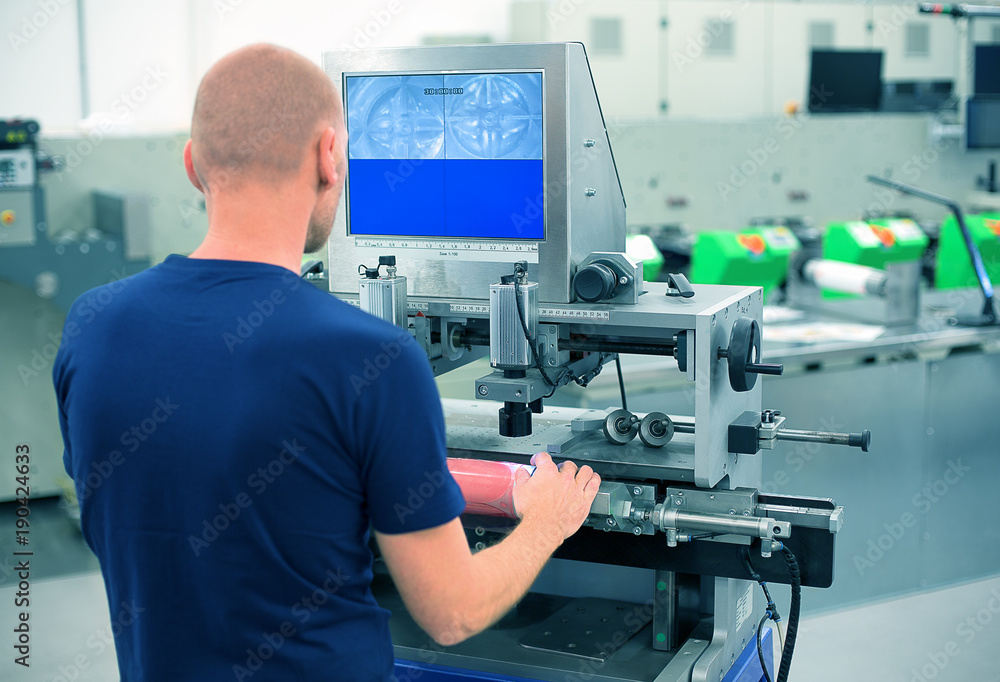 Worker in a printing and press center uses plate mounting machine to attach polymer relief plate on a printing cylinder. Scene showing mounting of printing plate for flexographic print machine. Worker in a printing and press center uses plate mounting machine to attach polymer relief plate on a printing cylinder. Scene showing mounting of printing plate for flexographic print machine.