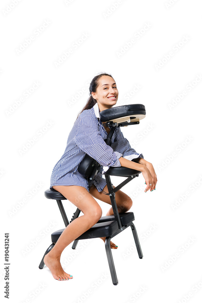 Young beautiful girl brunette office worker in a shirt and with bare legs sitting on a massage chair. Dark skin and hair of a brunette. Business concept and massage Young beautiful girl brunette office worker in a shirt and with bare legs sitting on a massage chair. Dark skin and hair of a brunette. Business concept and massage