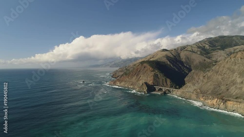 A static aerial shot of beautiful California coastline in summer weather. Bridge below with car crossing.