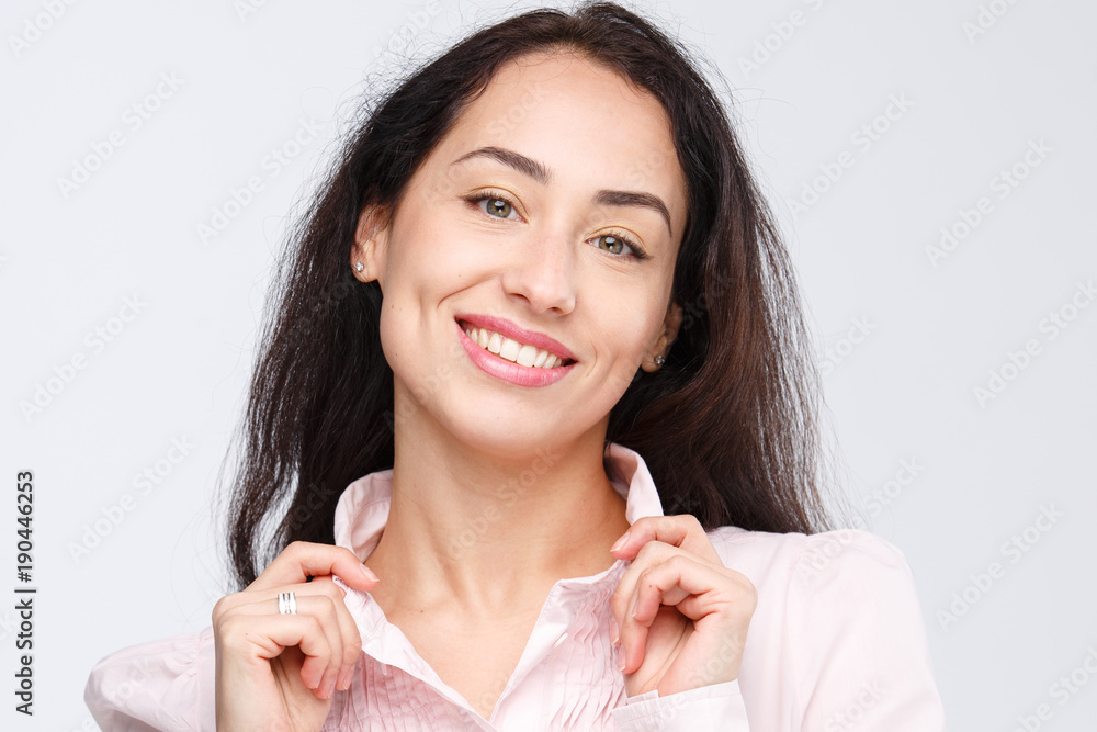 Close-up portrait of a young very beautiful woman with a charming toothy smile, black hair and brown eyes on a white background in a pink shirt. Hands near the head. Positive and joyful emotions Close-up portrait of a young very beautiful woman with a charming toothy smile, black hair and brown eyes on a white background in a pink shirt. Hands near the head. Positive and joyful emotions