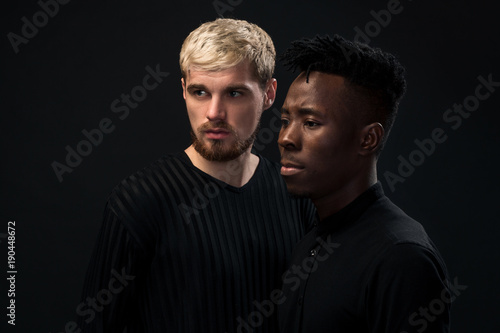 Portrait of two young african american and caucasian men standing over black background. Studio shot