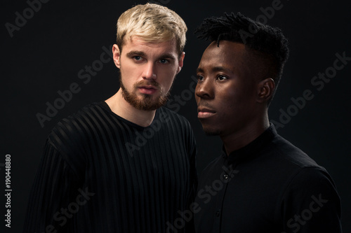 Portrait of two young african american and caucasian men standing over black background. Studio shot