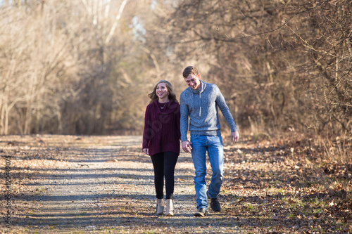 Young caucasian couple walking on dim gravel, dirt, rural, country road holding hands, kissing,smiling, laughing in love having fun season late winter bare trees © Mitch