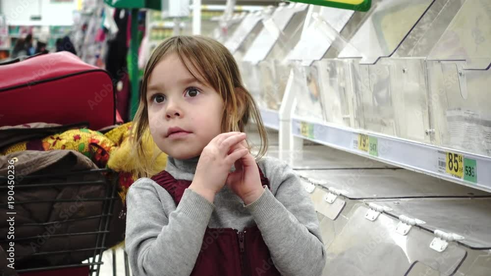 Little pretty girl in the supermarket. She has blonde hair, she is a blonde of European or American appearance, 4 - 5 years old. She wears winter clothes. The child looks at the goods. Little pretty girl in the supermarket. She has blonde hair, she is a blonde of European or American appearance, 4 - 5 years old. She wears winter clothes. The child looks at the goods.