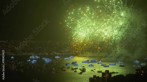 A wide shot of fireworks during New year's eve. Fireworks are being lit simultaneously over the harbour with neon lit boats and yachts floating beneath.