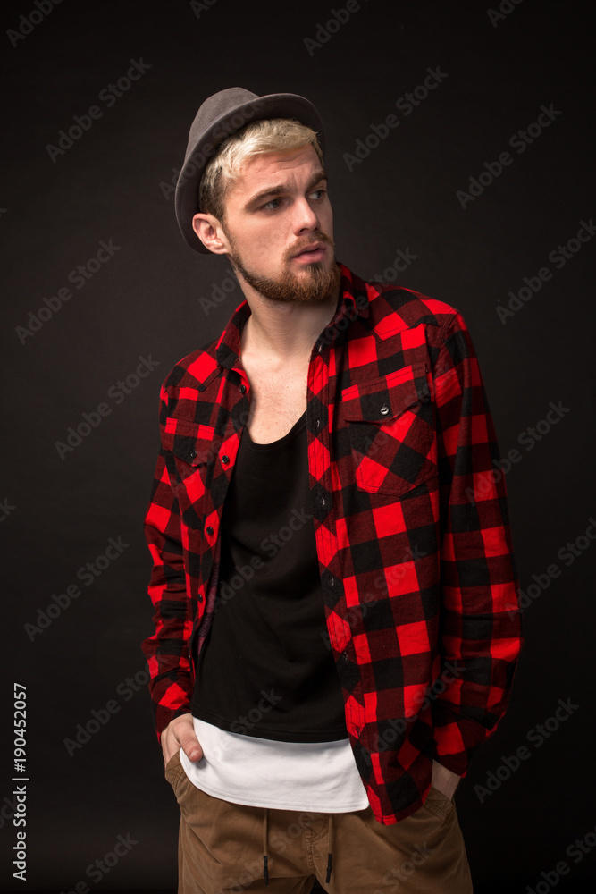 Handsome trendy young guy at the studio on black background. He wears beard and a plaid shirt
