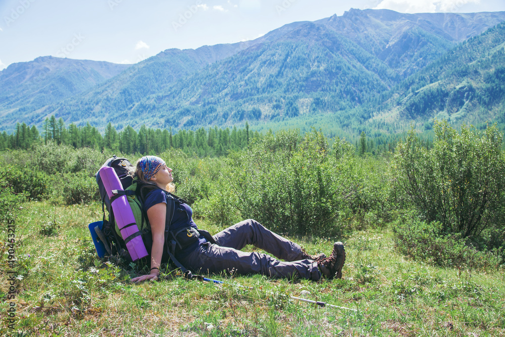 Woman hiker with backpack sitting on green grass and feel relax with closed eyes at background of high mountains