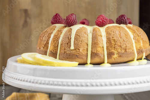 Lemon cake frosted with yellow sugar icing and red raspberries, dressed with lemon slices on the side on a white platter