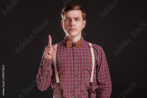 Attention! Strict Handsome young man in vintage shirt bow tie keeping finger raised and looking at camera while standing against black or grey background. People Emotions Fashion Businness Education photo