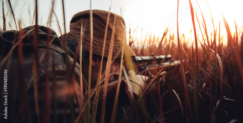 Hunter with rifle laying in long grass and concentrating on shooting prey