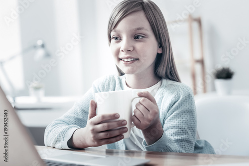 Cozy leisure time. Charming little girl focusing her attention on a screen of a laptop while sitting at a table and warming up with a cup of warm tea.