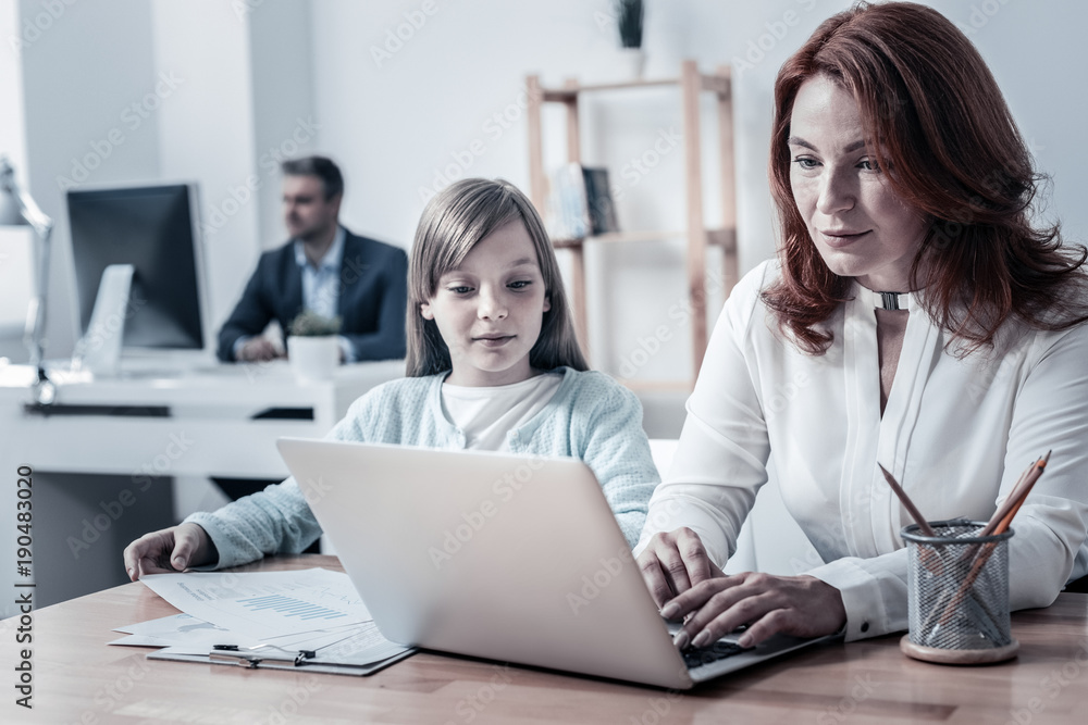 Working routine. Calm mother and daughter sitting next to each other and focusing their attention on a screen of laptop while spending their day at work. Working routine. Calm mother and daughter sitting next to each other and focusing their attention on a screen of laptop while spending their day at work.