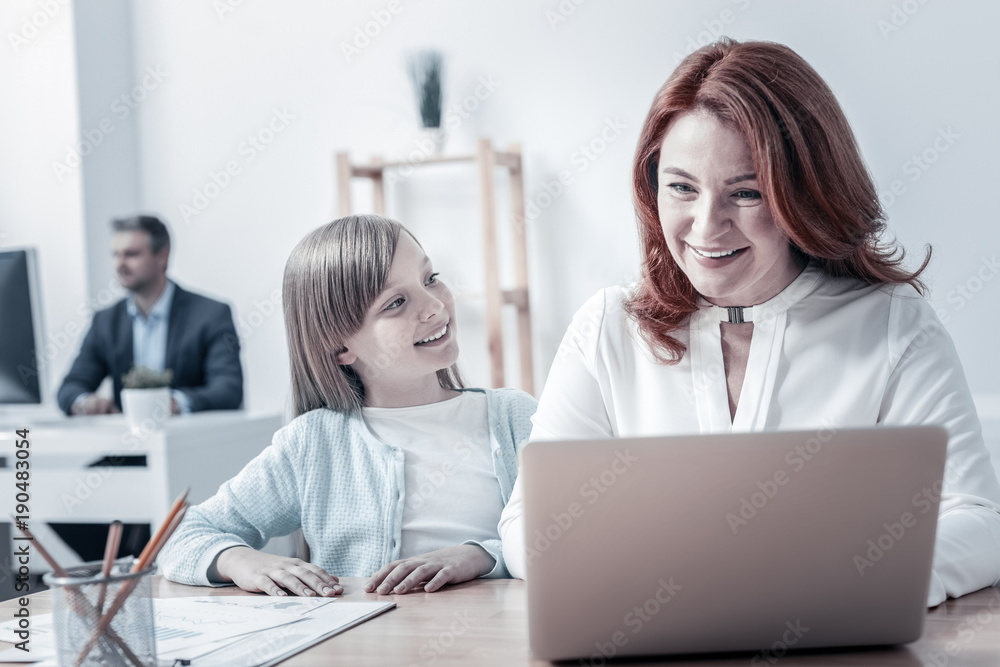 Pleasant day. Joyful mature woman smiling while working on a laptop and listening to the questions of her curious little daughter. Pleasant day. Joyful mature woman smiling while working on a laptop and listening to the questions of her curious little daughter.