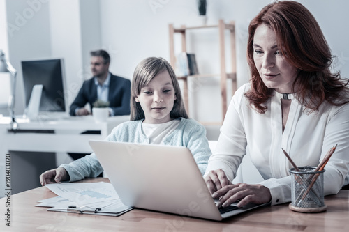Working routine. Calm mother and daughter sitting next to each other and focusing their attention on a screen of laptop while spending their day at work.