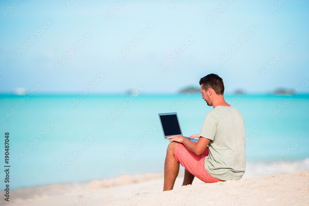 Young man sitting on sand with laptop on tropical caribbean beach. Man with computer and working on the beach