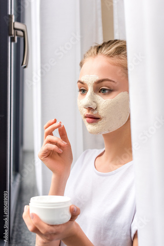 attractive young woman applying white clay mask on face at home
