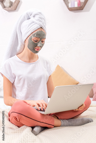 young woman with cosmetic clay mask working with laptop at home