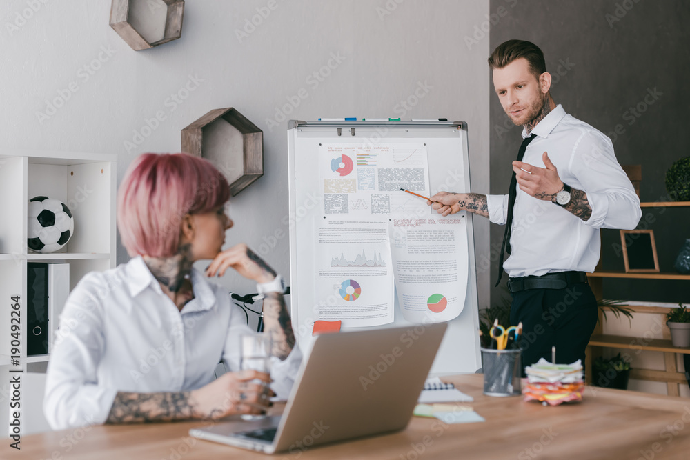 young businesswoman using laptop and looking at male colleague pointing at whiteboard with charts young businesswoman using laptop and looking at male colleague pointing at whiteboard with charts