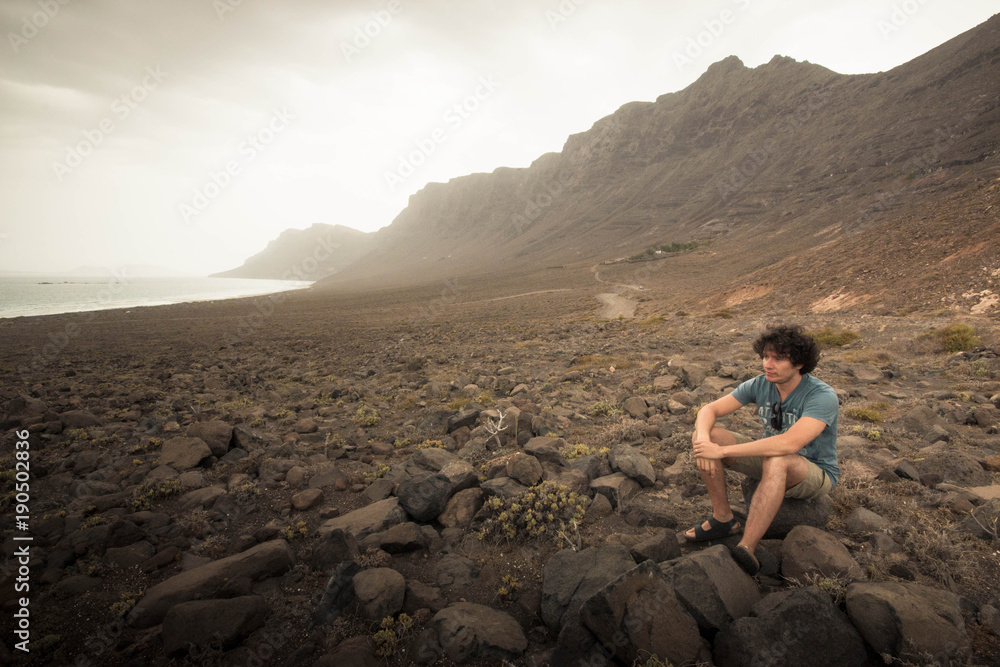 Fototapeta premium Young man sitting alone on a rock in Famara Beach, Lanzarote, Canary Islands. With copyspace.