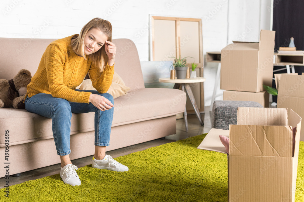 smiling young woman sitting on sofa while packing cardboard boxes during relocation smiling young woman sitting on sofa while packing cardboard boxes during relocation
