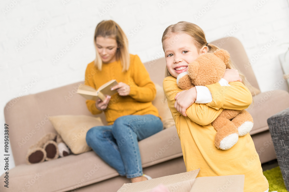 cute little girl hugging teddy bear and smiling at camera while mother reading book on sofa cute little girl hugging teddy bear and smiling at camera while mother reading book on sofa