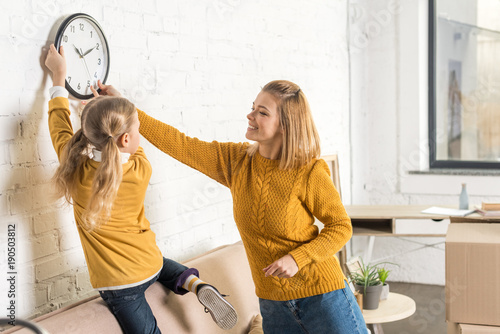 happy mother and daughter hanging clock on wall during relocation