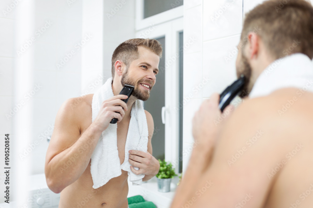 Handsome bearded man trimming his beard with a trimmer - obrazy, fototapety, plakaty 
