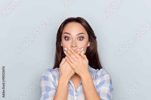 Close up portrait of attractive, pretty, cute, charming, woman closed her mouth with crossed hands and wide opened eyes, looking at camera, she can't believe her eyes, standing over grey background