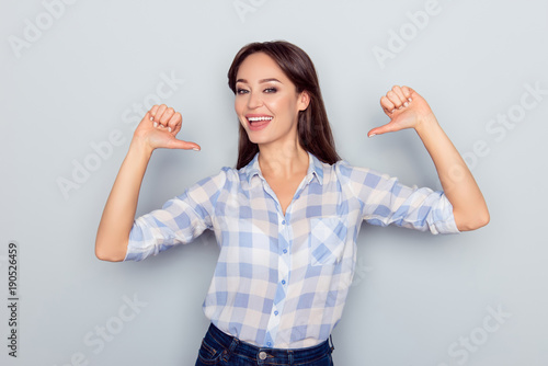 It is me! Portrait of smiling cheerful nice young woman in checkered shirt pointing thumb fingers at her body isolated on grey background, proud of herself, having ego