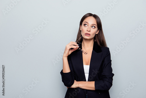 Portrait of young, nice, cute, perfect teacher holding eye wear in hand, biting eyelet, looking at copy space with thoughtful expression over grey background