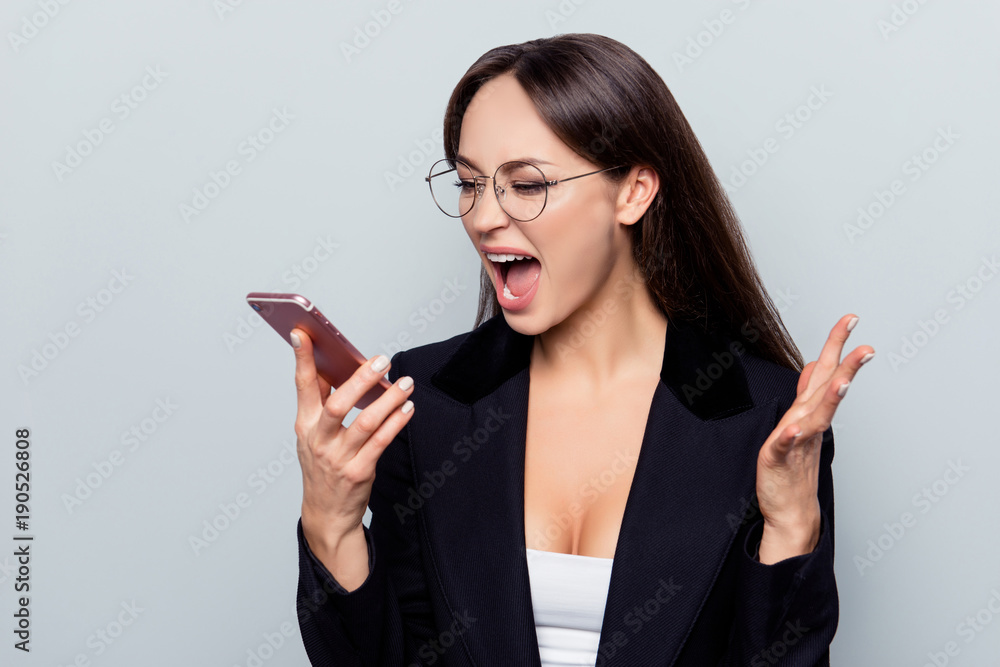 Portrait of frustrated, mad, crazy employer in formal wear, blach jacket yelling into smart phone while chatting with clients, gesturing with hand, she is out of herself, standing over grey background Portrait of frustrated, mad, crazy employer in formal wear, blach jacket yelling into smart phone while chatting with clients, gesturing with hand, she is out of herself, standing over grey background