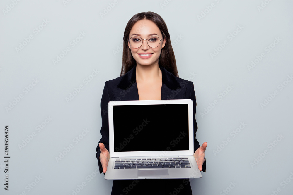 Portrait with copy space of pretty, cheerful, confident, professional, elegant, brunette in jacket demonstrating black screen and keypad of open laptop, standing over grey background Portrait with copy space of pretty, cheerful, confident, professional, elegant, brunette in jacket demonstrating black screen and keypad of open laptop, standing over grey background