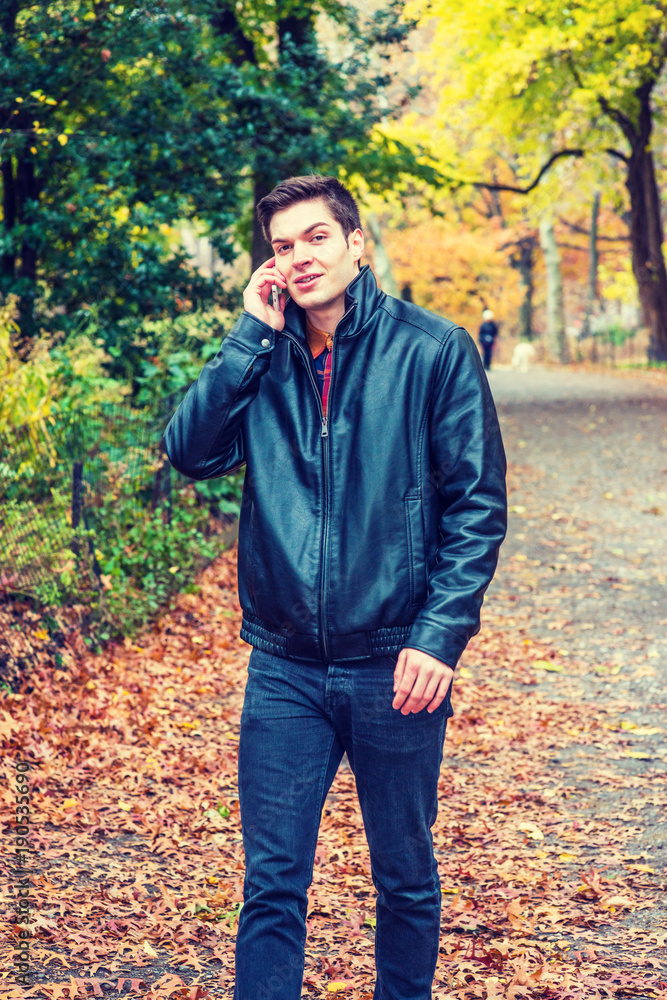 Young American man talking on cell phone, traveling at Central Park, New York in autumn day, wearing black leather jacket, jeans, walking on road with colorful trees, fallen leaves, smiling..