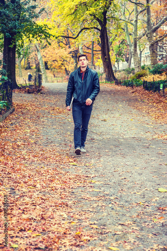 Young American man traveling at Central Park, New York in autumn day. Man wearing black leather jacket, jeans, gray casual shoes, holding laptop computer, walking on road with colorful trees, leaves