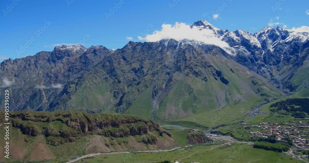 Village on snow-capped peaks of mountains Background In Kazbegi District, Military-Georgian road. Georgia. Panoramic Landscape At Spring Or Summer Season. Village on snow-capped peaks of mountains Background In Kazbegi District, Military-Georgian road. Georgia. Panoramic Landscape At Spring Or Summer Season.