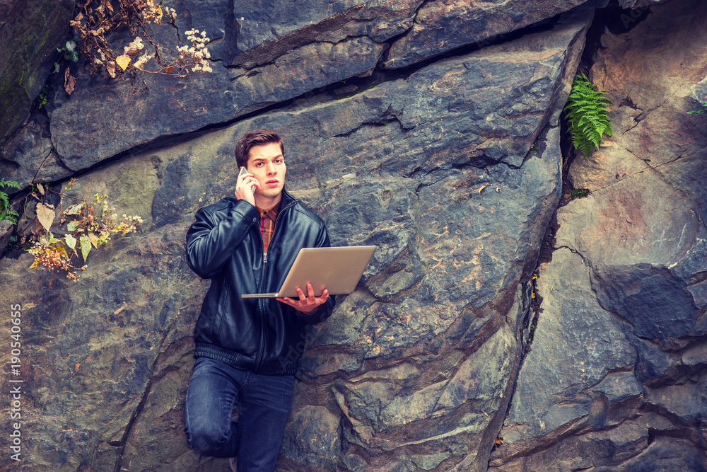 Young American Man wearing black leather jacket, pants, standing against rocks at Central Park, New York, working on laptop computer, talking on cell phone.. Young American Man wearing black leather jacket, pants, standing against rocks at Central Park, New York, working on laptop computer, talking on cell phone..