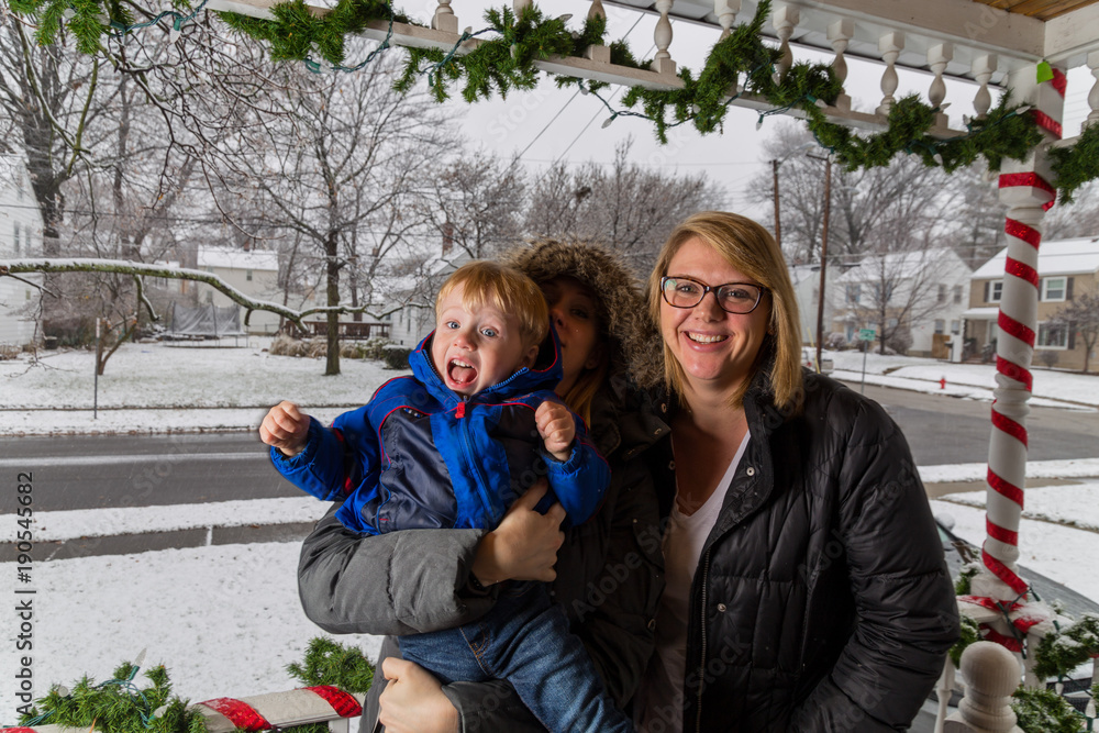 Two Caucasian Women And A Young Toddler Boy Stand On A Front Porch As It Snows Looking At The Christmas Decorations. Two Caucasian Women And A Young Toddler Boy Stand On A Front Porch As It Snows Looking At The Christmas Decorations.