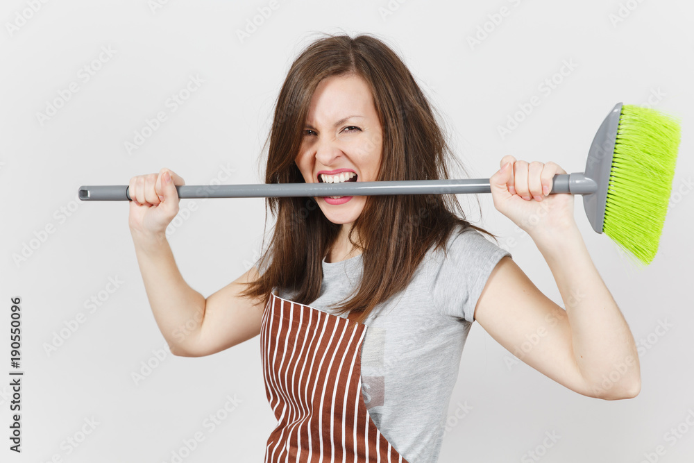 Young fun crazy dizzy loony wild aggressive housewife with brown tousled hair in striped apron isolated on white background. Mad witch woman holding, biting teeth broom. Copy space for advertisement. Young fun crazy dizzy loony wild aggressive housewife with brown tousled hair in striped apron isolated on white background. Mad witch woman holding, biting teeth broom. Copy space for advertisement.