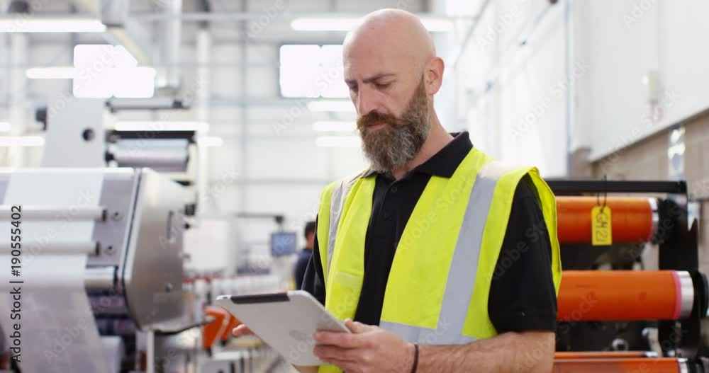 4K Worker in printing factory looking down at computer tablet with machinery in background 4K Worker in printing factory looking down at computer tablet with machinery in background