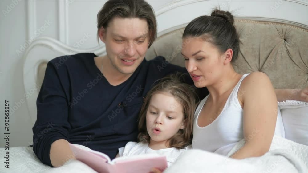 Clever Little Girl is Reading Aloud a Book Lying on the Bed. Her Young Parents are Helping Her. Female Child is Learning Foreing Language with Family. Clever Little Girl is Reading Aloud a Book Lying on the Bed. Her Young Parents are Helping Her. Female Child is Learning Foreing Language with Family.