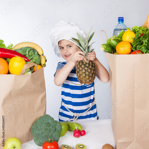 Little happy girl in chef hat with big bags of products holding a pineapple. A variety of fresh fruits and vegetables in bags on the table. photo