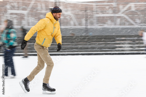 Full length portrait of bearded man in yellow jacket, beige trousers, black hat on ice rink, outdoors in snowy winter day/ Weekend activities outdoor in cold weather/man shooting in motion