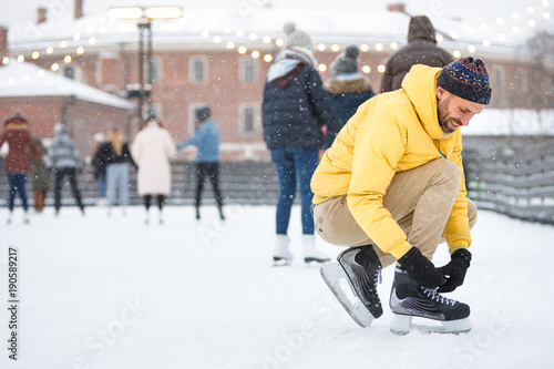 Close up bearded man in yellow jacket, beige trousers and wearing black skates on ice rink /Man tying black skates on ice rink in snowy winter day /Weekends activities in cold weather/