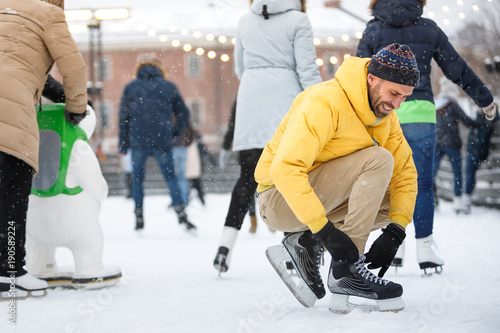Close up handsome bearded man in yellow jacket, and beige trousers, laces up black skates on ice rink/ Man tying black skates on ice rink in snowy winter day/ Weekends activities in cold weather