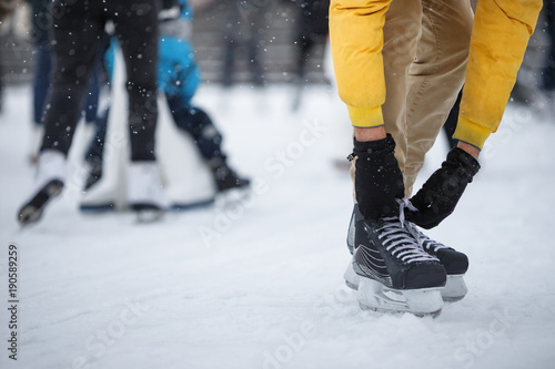 Close up of man hands wearing black skates on ice rink /Man tying black skates on ice rink in snowy winter day, people on background /Weekends activities in cold weather/ Winter time concept